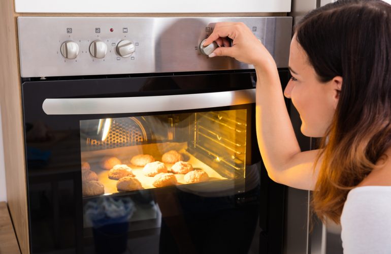 Young Woman Using Microwave Oven For Baking Fresh Cookies In Kitchen